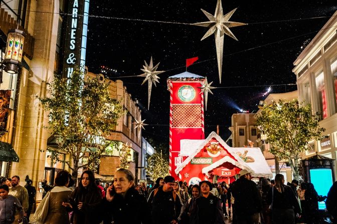 Festive night in a city shopping district with crowds strolling under hanging star lights and faux snow by a giant red gingerbread clock tower and illuminated trees.