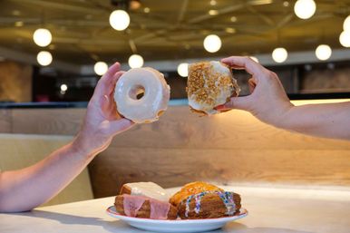 Two hands hold up glazed donuts — a classic ring and a crumb-topped variety — above a plate of assorted iced pastries on a cafe table with wooden booth seating and round pendant lights.
