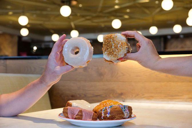 Two hands hold up glazed donuts — a classic ring and a crumb-topped variety — above a plate of assorted iced pastries on a cafe table with wooden booth seating and round pendant lights.