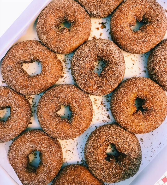 Top-down view of cinnamon-sugar donuts in a white bakery box — golden-brown, sugar-crusted breakfast treats with scattered crumbs