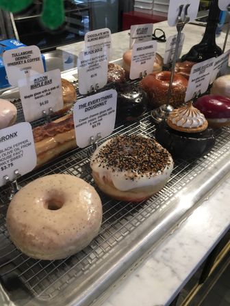 Assorted artisan doughnuts on a wire rack in a bakery display case on a marble counter — glazed, chocolate, bacon-maple and an Everything doughnut topped with sesame and poppy seeds.