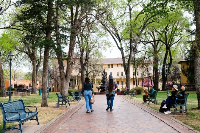 Couple strolling down a tree-lined brick promenade toward a central fountain in a sunny urban park plaza with green benches and people relaxing