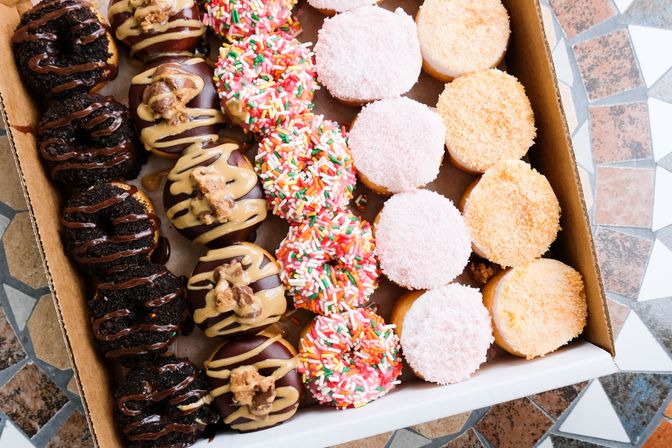 Box of assorted mini donuts on a mosaic tile table: chocolate cookie, peanut-butter-drizzled, colorful sprinkle, pink coconut and orange crumb-topped varieties