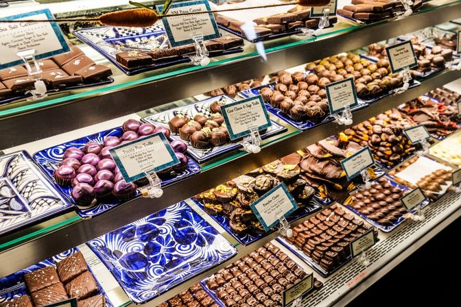 Glass display of artisan chocolates and truffles on blue ceramic trays in a chocolate shop — glossy purple heart truffles, pistachio clusters, salted chocolate squares and small labeled cards.