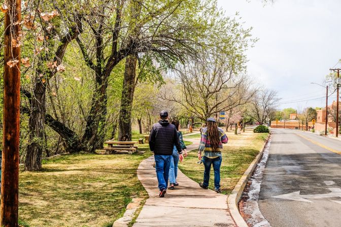 Three people walking along a winding, tree-lined park sidewalk beside a quiet suburban road in early spring.