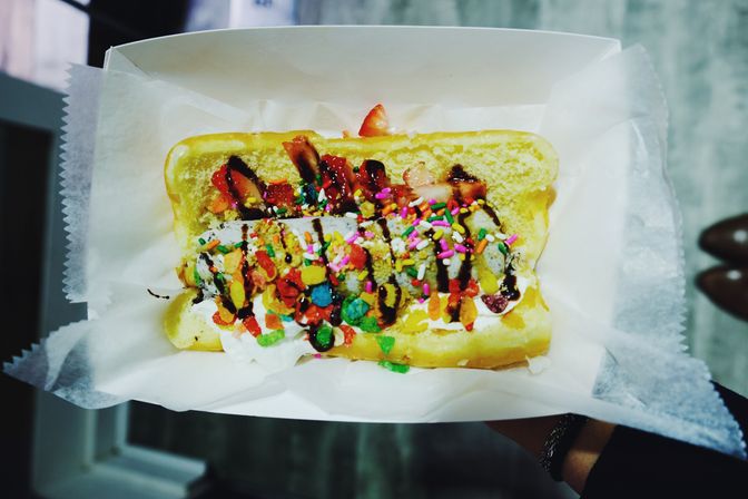 Top-down view of a dessert hot dog: sweet split bun filled with ice cream and whipped cream, sliced strawberries, chocolate drizzle, rainbow sprinkles and crunchy colorful cereal in a paper tray.