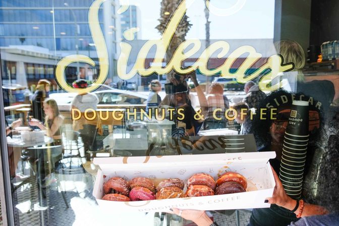 Box of assorted colorful doughnuts held up to a sunlit downtown café window reading "Doughnuts & Coffee", patrons and street reflections visible inside