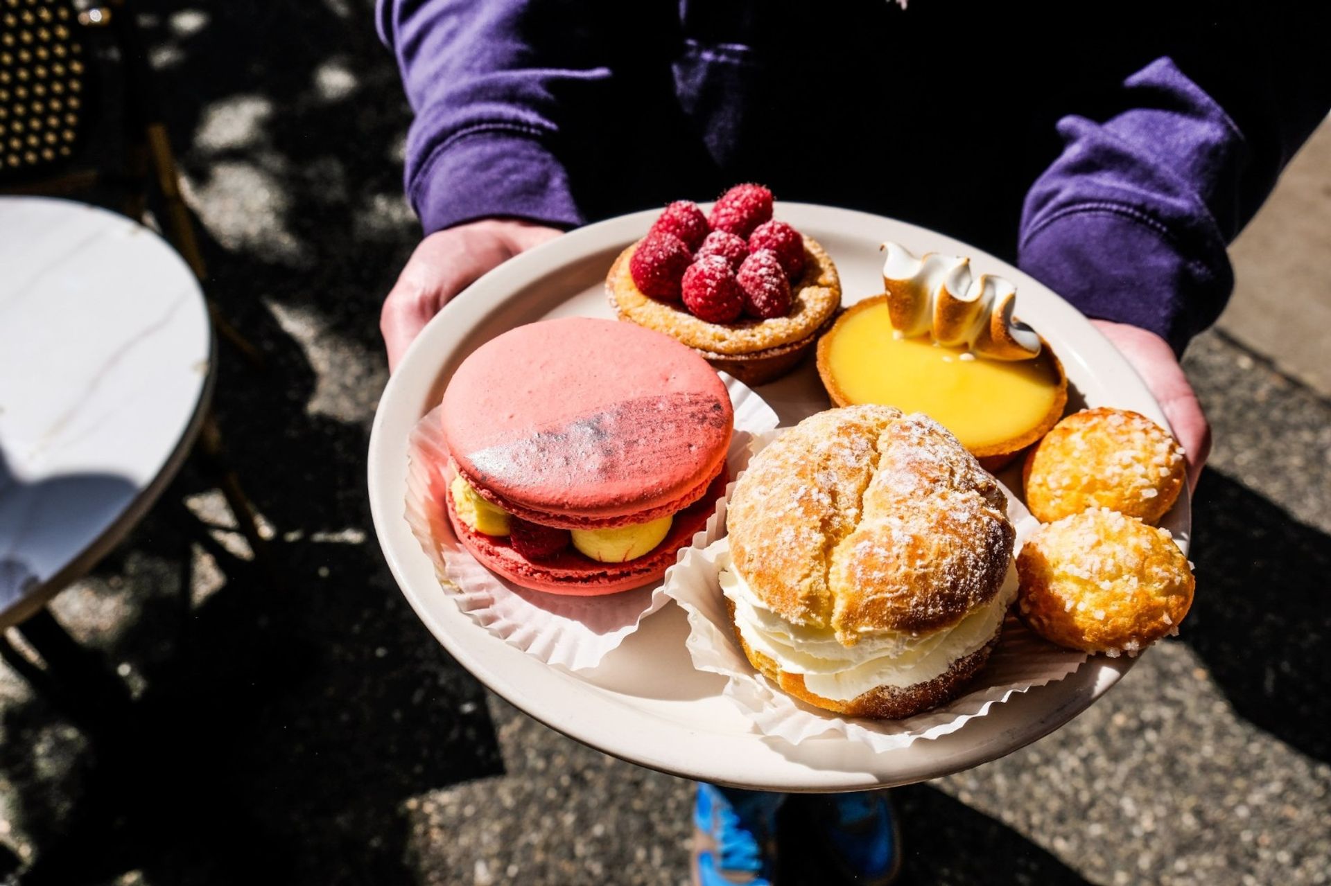 Plate of assorted pastries at an outdoor cafe patio: large pink macaron filled with cream and raspberries, raspberry tartlet, lemon tart with toasted meringue, cream-filled choux and two mini cream puffs.