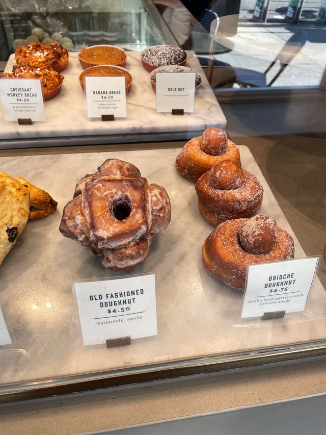 Bakery glass display with artisan pastries on marble trays—glazed old-fashioned doughnut, sugar‑coated brioche doughnuts, banana bread and croissant monkey bread visible with small price cards.