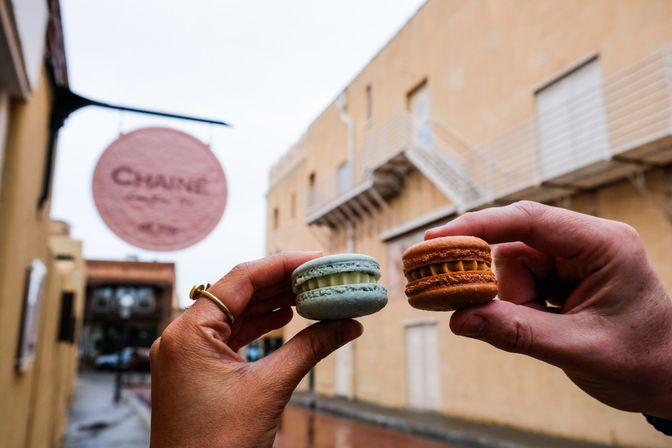 Two hands holding a pastel blue and an orange macaron in a rainy downtown alley with beige buildings and a blurred round bakery sign in the background
