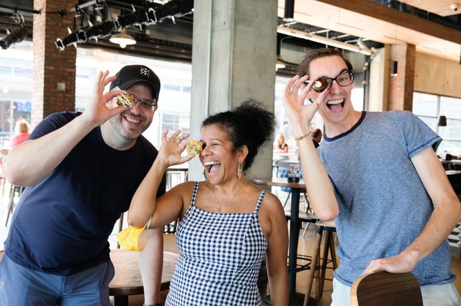 Three friends laughing in a bright downtown café food hall, playfully holding colorful mini donuts up to their eyes
