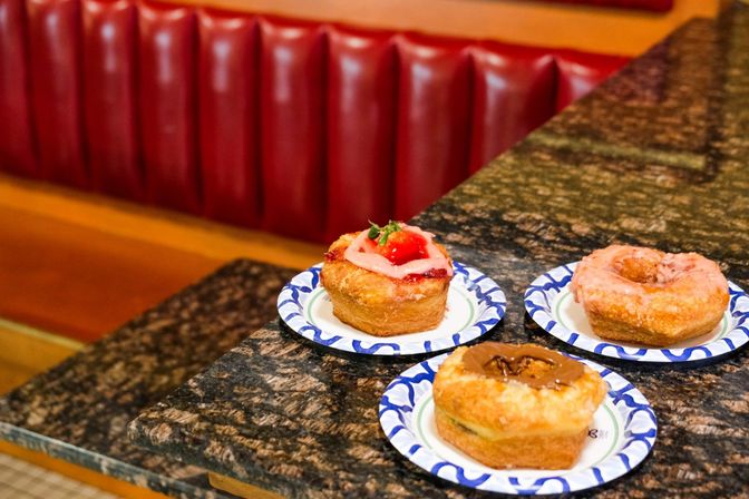 Three assorted donuts—one with fresh strawberry and pink icing, one glazed, and one with chocolate topping—on paper plates on a marble diner counter in front of a red vinyl booth.