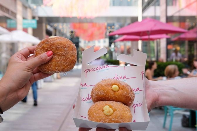 Close-up of hands holding sugar-coated custard-filled doughnuts in a white takeout box at an outdoor mall café with pink umbrellas and blurred shoppers in a city plaza.
