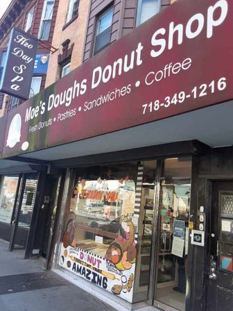 City sidewalk donut shop storefront with maroon awning, glass door and display window painted with colorful donut characters and pastry artwork advertising fresh donuts, coffee and pastries.