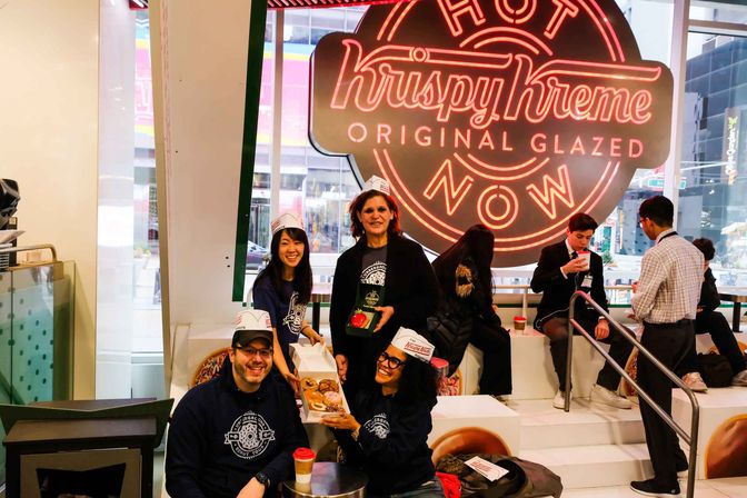 Smiling group in paper baker hats inside a bright downtown donut shop with a large neon glazed-donut sign, holding boxes of donuts and coffee by window seating