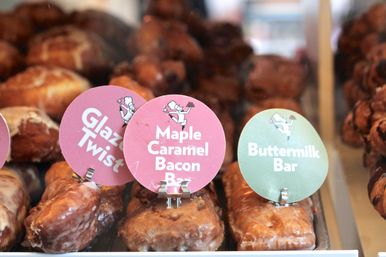 Glass bakery case with golden-brown glazed pastry bars and colorful round labels reading "Maple Caramel Bacon Bar", "Buttermilk Bar", and "Glazed Twist".