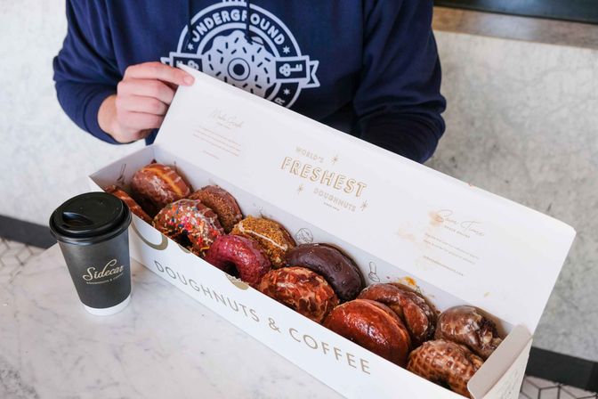 Assorted colorful gourmet doughnuts with glazes and sprinkles in a long white box labeled doughnuts & coffee, next to a to‑go coffee cup on a marble cafe table as a person opens the box.