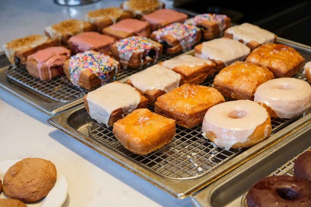 Fresh-baked assorted glazed and frosted square cronuts and ring donuts on wire racks at a bakery display, featuring sprinkled, pink, vanilla-glazed and salted caramel varieties.