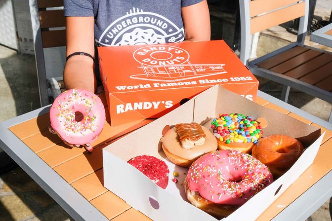 Person’s arm holding a pink sprinkled donut next to a bright orange donut box and an open box of assorted glazed, caramel and candy‑topped donuts on a sunny outdoor cafe table.