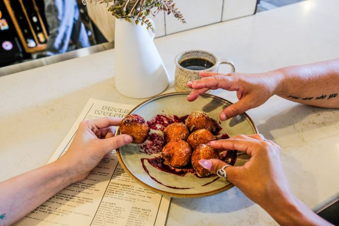 Close-up of hands grabbing sugared donut holes with berry sauce on a ceramic plate at a bright brunch cafe counter, with a coffee mug, menu and small vase nearby.