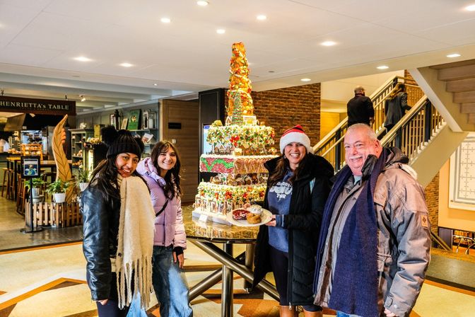 Four smiling people in winter coats pose in a hotel lobby beside a towering, multi-tiered holiday gingerbread display; one wears a Santa hat and holds a plate of pastries.