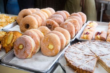 Bakery display of sugar-coated custard- and jam-filled doughnuts on metal trays, alongside fruit pastries and powdered-sugar tart slices