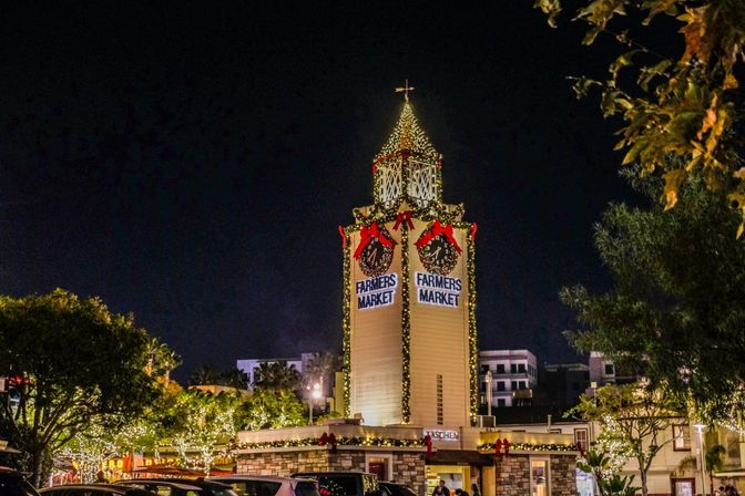 Festive clock tower glowing at night above an outdoor market, draped in twinkling holiday lights, wreaths, and red bows.
