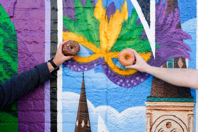 Two hands hold glazed donuts up to the eye holes of a colorful Mardi Gras-style mask mural painted on a brick wall, a festive street art scene