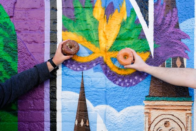 Two hands hold glazed donuts up to the eye holes of a colorful Mardi Gras-style mask mural painted on a brick wall, a festive street art scene