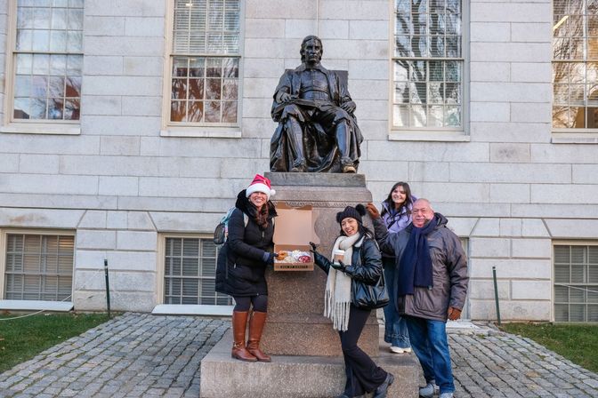 Four visitors in winter coats smiling and posing by a seated bronze statue on a historic college campus; one wearing a Santa hat holds an open box of donuts and another holds coffee.