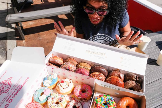 Excited person leaning over boxes of colorful assorted doughnuts with sprinkles and toppings on an outdoor table.