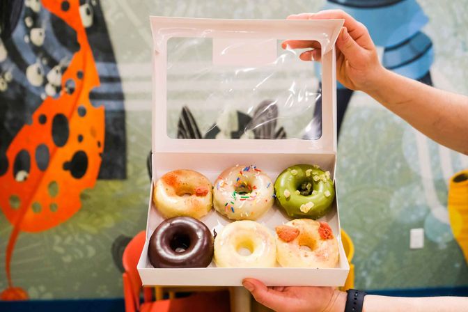Hands holding an open white box of six assorted colorful mini donuts—chocolate, matcha, glazed with sprinkles and fruit pieces—against a bright café mural background.
