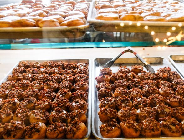 Rows of fresh glazed mini donuts topped with crispy bacon crumble on metal trays in a bakery display case