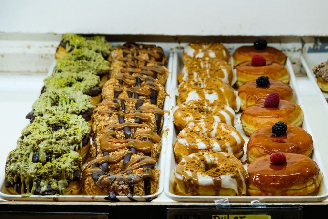 Assorted gourmet donuts in a bakery display case: pistachio‑drizzled chocolate, caramel‑and‑chocolate coconut, white‑glazed cinnamon, and caramel‑glazed donuts topped with raspberries and blackberries.