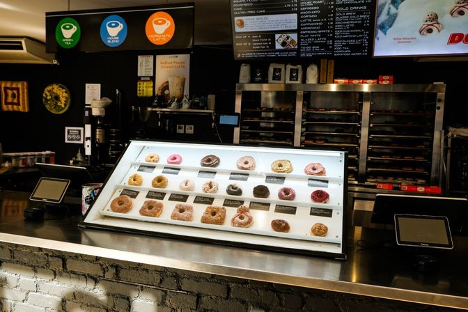 Assorted donuts in a lit glass display case on a coffee shop counter, with menu boards above, POS terminals and baking racks in the background.