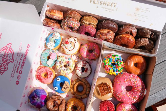 Assorted colorful gourmet donuts in open bakery boxes on a wooden table — glazed, sprinkled, cereal- and candy-topped, cookie‑monster and floral-decorated varieties perfect for brunch or a party.