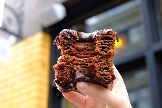 Hand holding a chocolate‑glazed cronut — flaky, layered croissant‑donut with rich chocolate filling against an urban café window backdrop