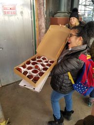 Smiling woman in a winter coat and backpack holds open a cardboard box of dozen glazed donuts in paper cups on an urban sidewalk under scaffolding as another person in a knit hat looks on.
