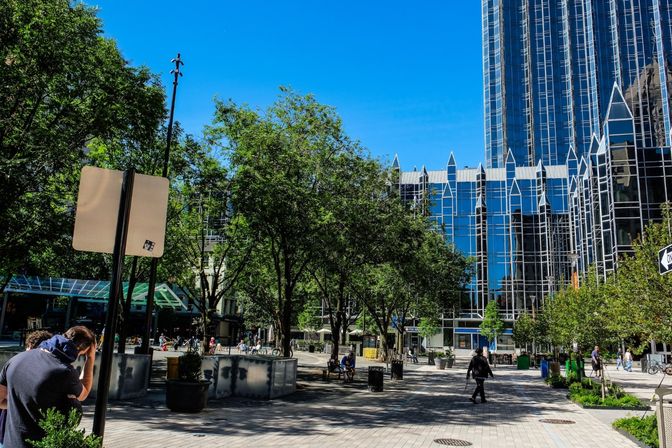 Sunny downtown urban plaza with shaded trees, benches and pedestrians on a paved walkway in front of a reflective modern glass skyscraper under a bright blue sky.