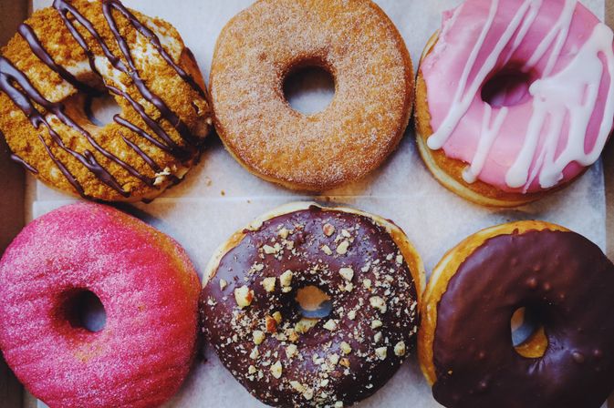 Top-down view of six assorted gourmet donuts in a box: crumb-topped with chocolate drizzle, cinnamon-sugar, pink glazed with white drizzle, bright pink sugar, chocolate with chopped nuts, and chocolate glazed.