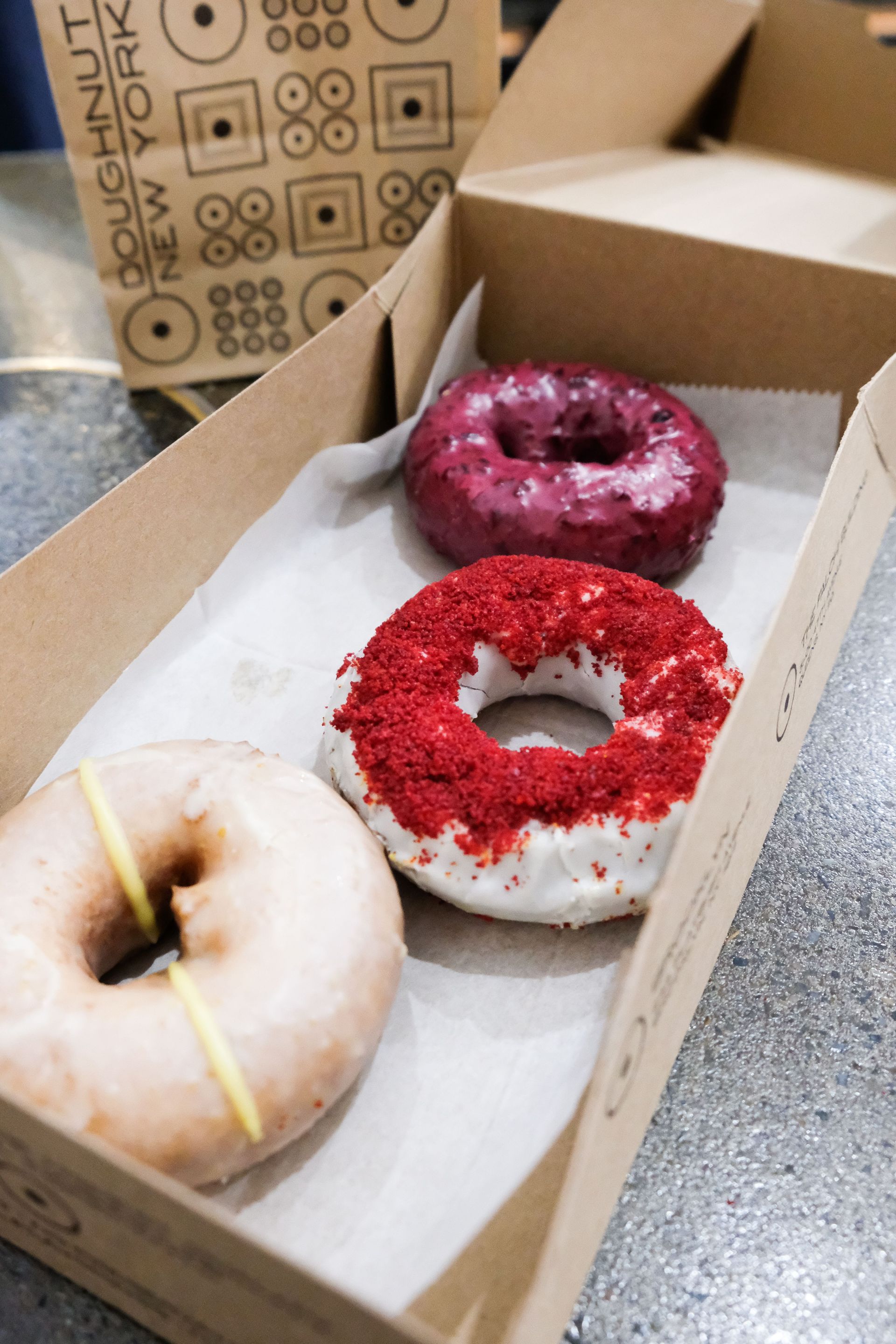 Three assorted colorful donuts — light glazed with a lemon stripe, white-frosted with red crumbs, and purple berry-glazed — in a cardboard takeout box on a cafe countertop