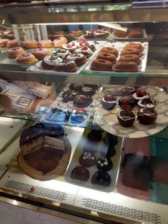 Glass bakery display case filled with assorted pastries: pink‑frosted and chocolate donuts, raspberry‑topped chocolate cupcakes, ring cakes, a sliced layered chocolate cake, and chocolate cookies on trays.