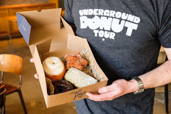 Assorted gourmet donuts in a cardboard box — cream-swirl, powdered, star-shaped and chocolate-glazed — held inside a cozy urban cafe by someone wearing a donut-themed t-shirt.