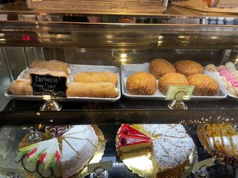 Bright bakery display case showcasing sugar-coated filled donuts, golden fried pastry rounds with crunchy coating, colorful macarons, and assorted powdered-sugar and fruit-topped cake slices.