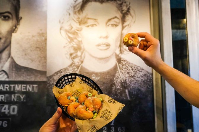 Hand holding a mini cereal-topped donut above a paper-lined basket of sugar-coated mini donuts with colorful cereal, set against a large black-and-white vintage celebrity mural in a trendy dessert cafe.