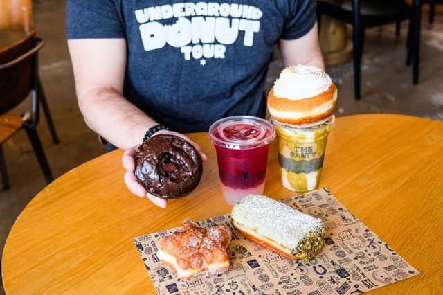 Person at a cafe holding a chocolate-glazed donut over a wooden table with assorted pastries — a sugar-dusted cruller, pistachio-coated filled donut, a layered berry drink and a creamy shake topped with a small donut.