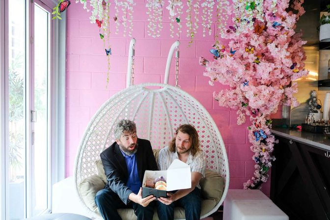 Two men sitting in a white hanging egg chair in an Instagrammable pink cafe nook with faux cherry blossoms and butterflies, opening a box of colorful pastries.