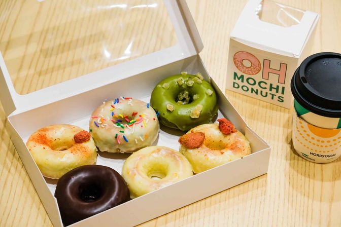 Assorted mini mochi donuts in a white takeout box on a light wooden table — matcha, chocolate, sprinkled and glazed varieties — served with a takeaway coffee cup