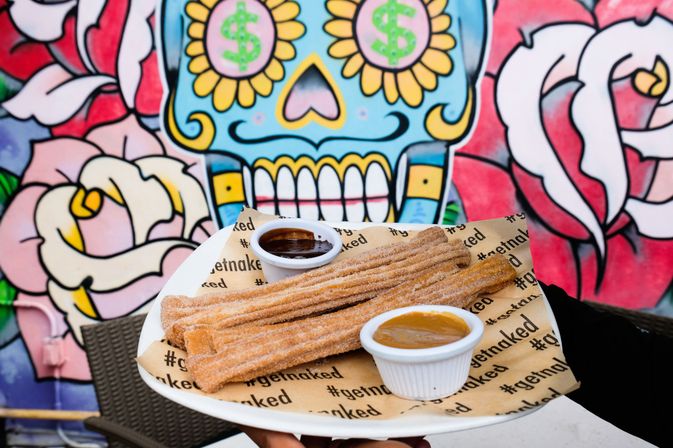 Crispy cinnamon-sugar churros with chocolate and caramel dipping sauces on a plate held in front of a vibrant Day of the Dead sugar skull street-art mural.