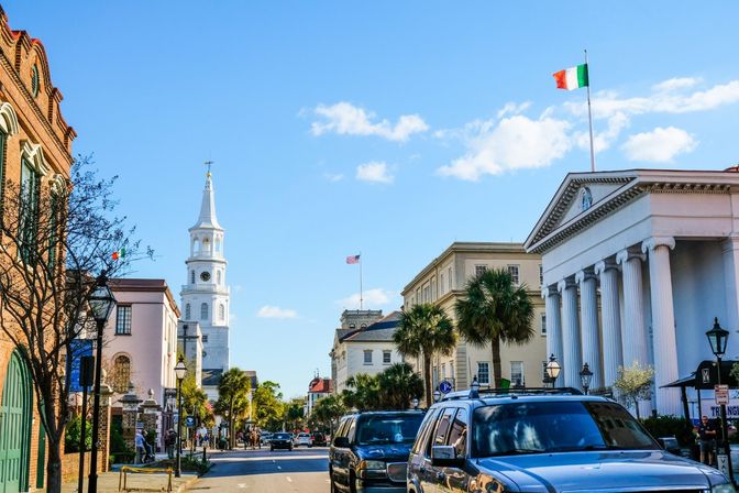 Sunny historic downtown street with a white church steeple, neoclassical columned building flying an Irish flag, palm trees lining the road and parked cars beneath a bright blue sky.